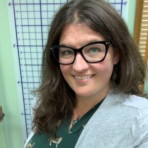 White woman with long brown hair and blue eyes, wearing heavy frame black glasses, a green dress, and a gray sweater. She is smiling.