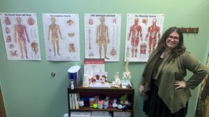 A woman standing with a shelf that contains several anatomic models. Anatomic posters are on the wall behind her